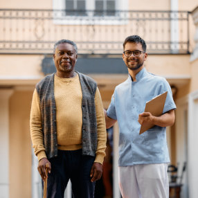 Smiling black senior man and young caregiver in front of residential care home looking at camera.